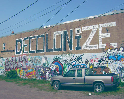 a wall is half covered in graffiti, in the top half, which is mostly unadorned, the word 'DECOLONIZE' is painted in large black and white letters